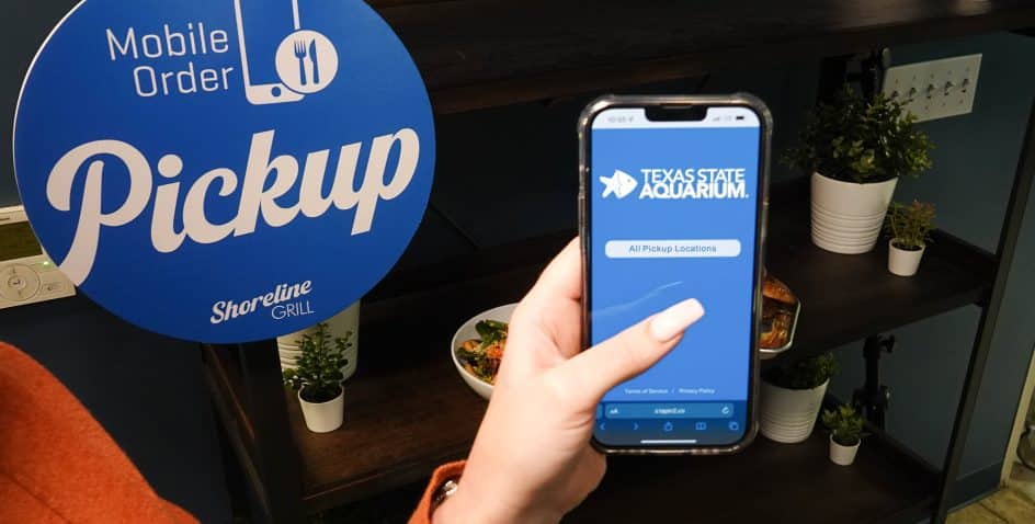Hand holding a phone showing the Texas State Aquarium's mobile food ordering page next to the Shoreline Grill's mobile order pickup station