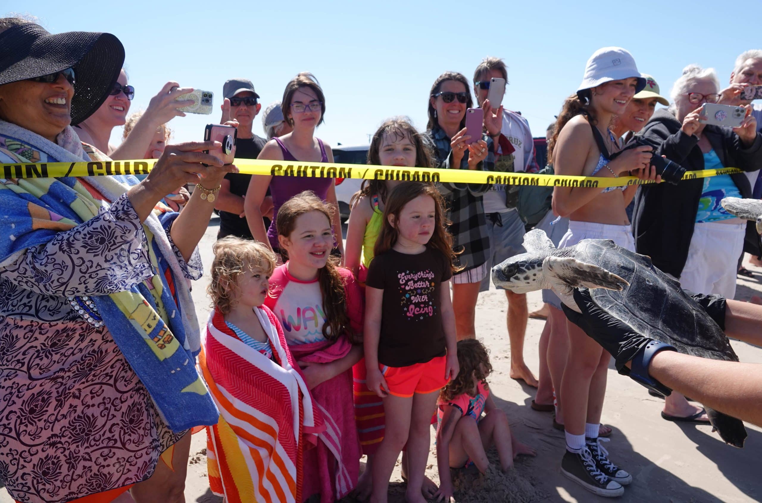 A group of excited people, including several children with towels, stand behind yellow caution tape on a sunny beach, watching gloved hands release a sea turtle toward the sand. Many are taking photos.