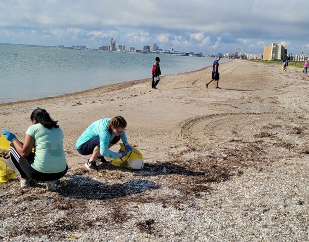 People wearing gloves pick up trash and collect it in large yellow bags on a sandy beach, with more volunteers cleaning in the distance. The sky is partly cloudy and buildings line the shore in the background.