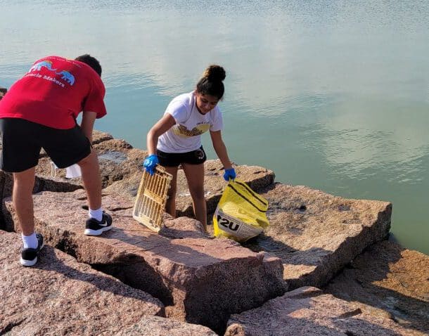 Two people clean up litter on large rocks by the water; one person holds a yellow trash bag and a piece of debris, while the other bends down to pick up trash. Both are wearing gloves.