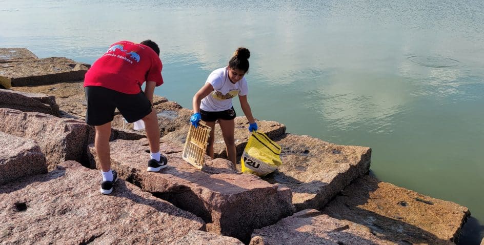 Two people clean up litter on large rocks by the water; one person holds a yellow trash bag and a piece of debris, while the other bends down to pick up trash. Both are wearing gloves.