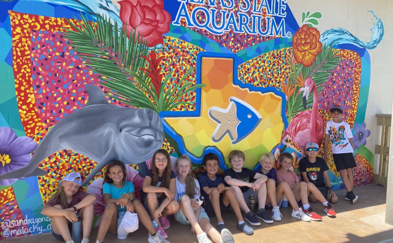 A group of smiling children sit in front of a colorful mural at the Texas State Aquarium, featuring a dolphin, fish, flowers, palm leaves, and a large orange Texas state outline.