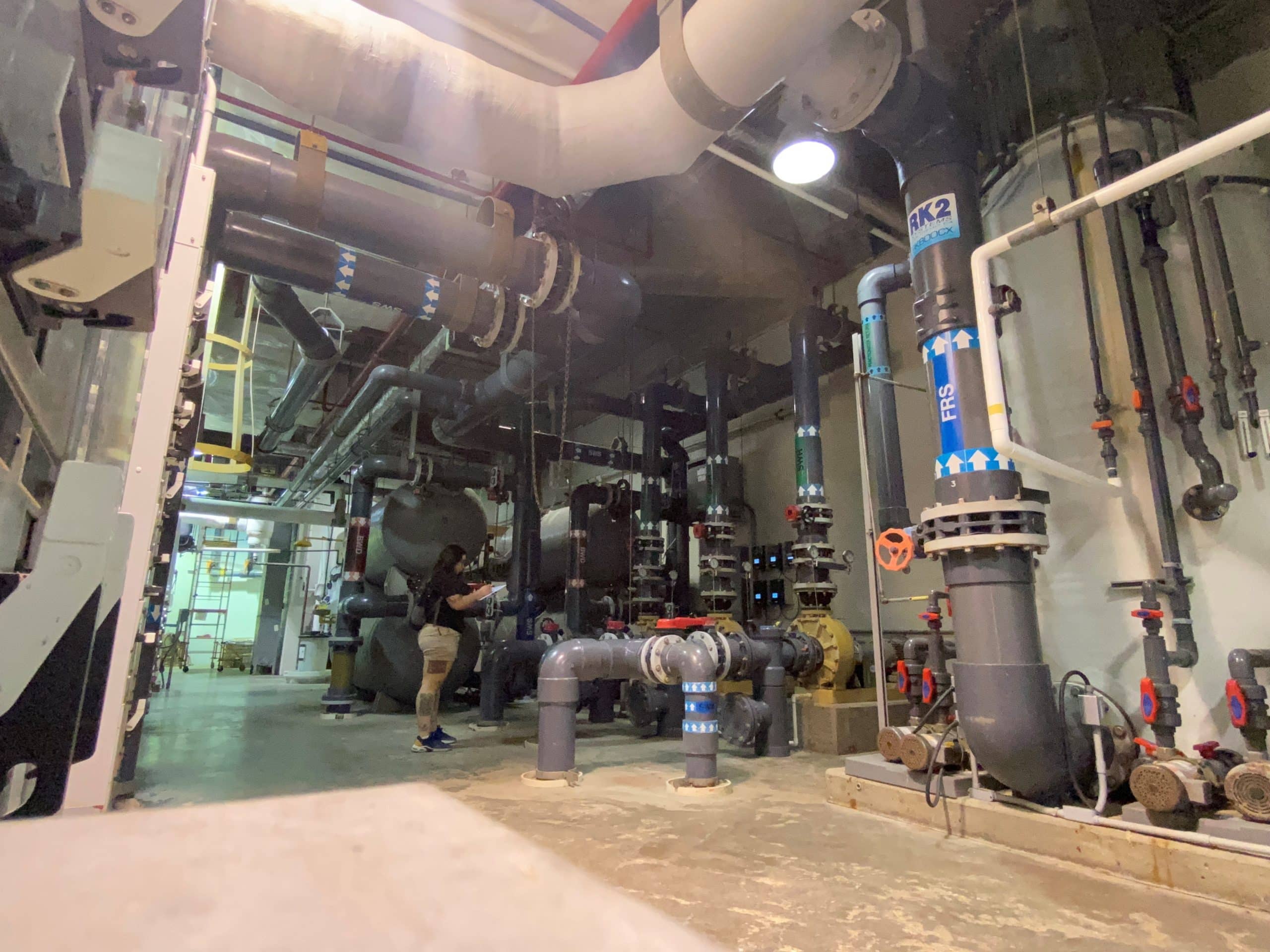 A person stands in a large industrial room filled with pipes, tanks, and valves. The setting appears to be a mechanical or boiler room with various equipment and overhead ductwork.