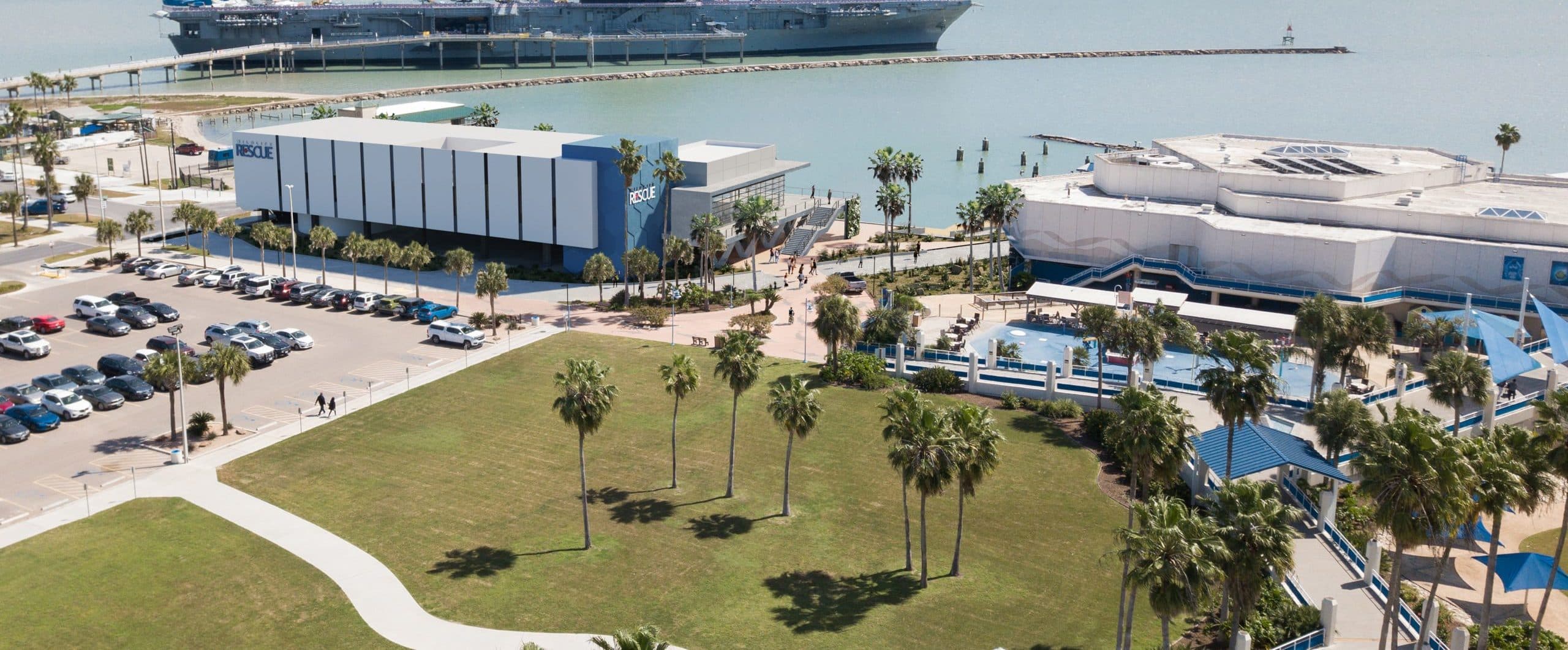Aerial view of the Texas State Aquarium with nearby green lawn, palm trees, parked cars, and the USS Lexington aircraft carrier docked on the water in Corpus Christi, Texas.