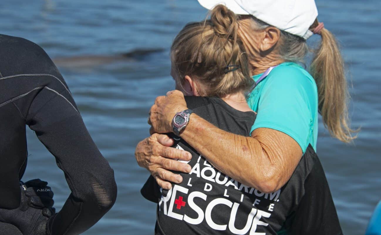 Two women, one in a Rescue shirt and the other in a blue shirt and white cap, hug near the water. Another person in a wetsuit stands nearby. They appear to be part of a wildlife rescue effort.