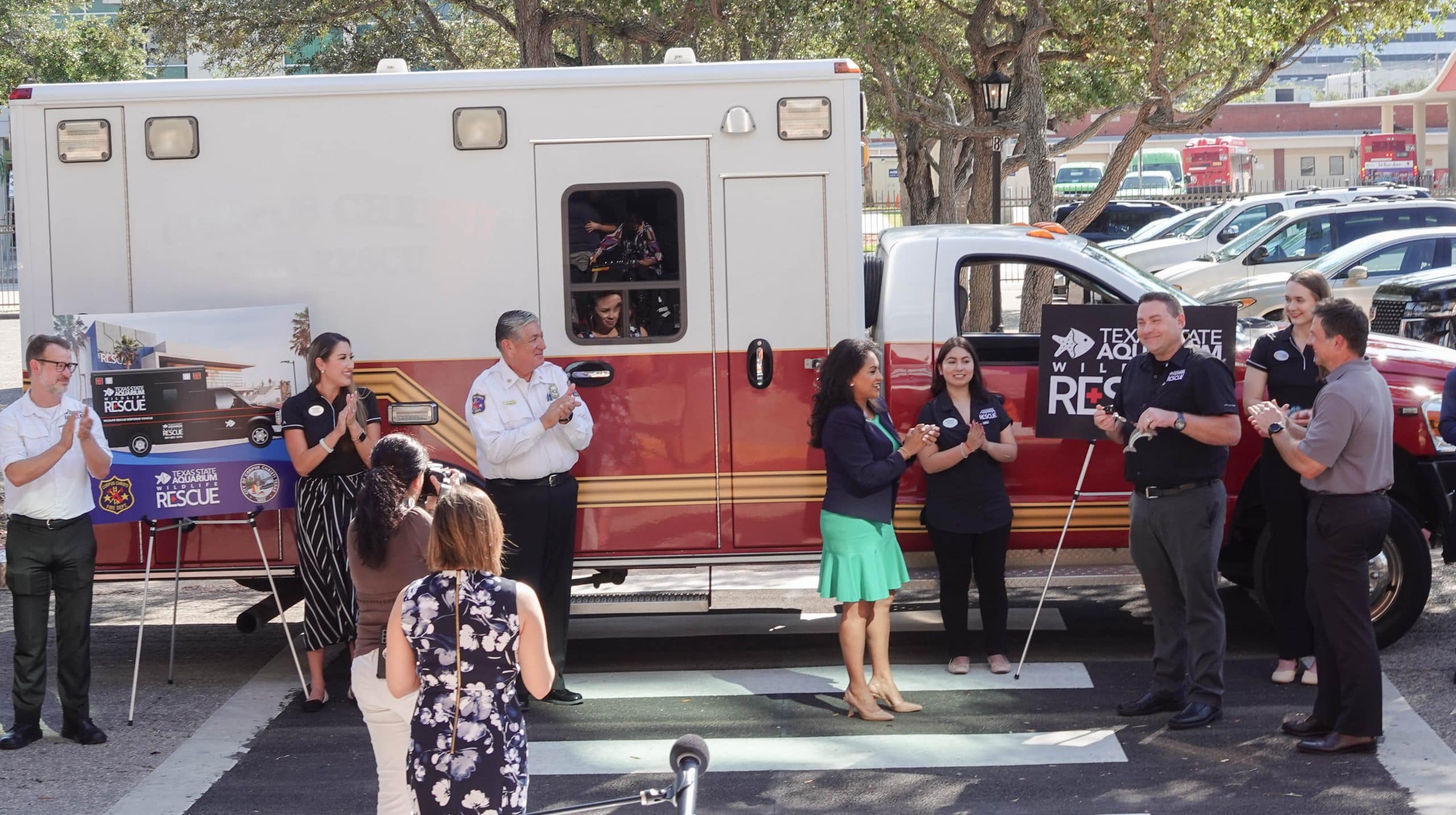 A group of people stand and talk in front of a Texas A&M wildland rescue vehicle during an outdoor event, with posters on display and several people holding white canes.