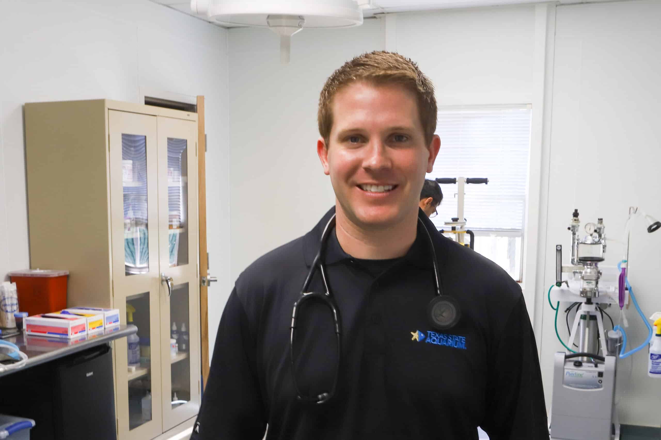 A man wearing a black shirt and a stethoscope around his neck smiles while standing in a brightly lit medical or veterinary exam room with equipment and cabinets in the background.