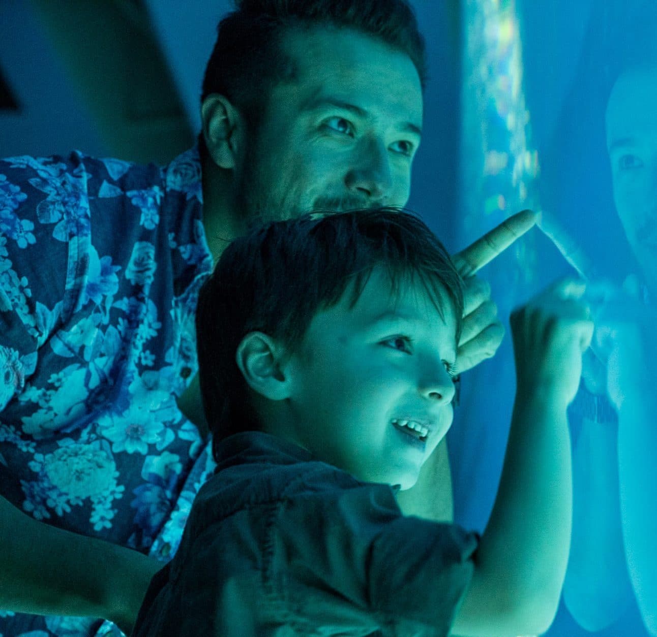 A smiling child and an adult, possibly a parent, point excitedly at something inside a blue-lit aquarium, their faces illuminated by the glow.