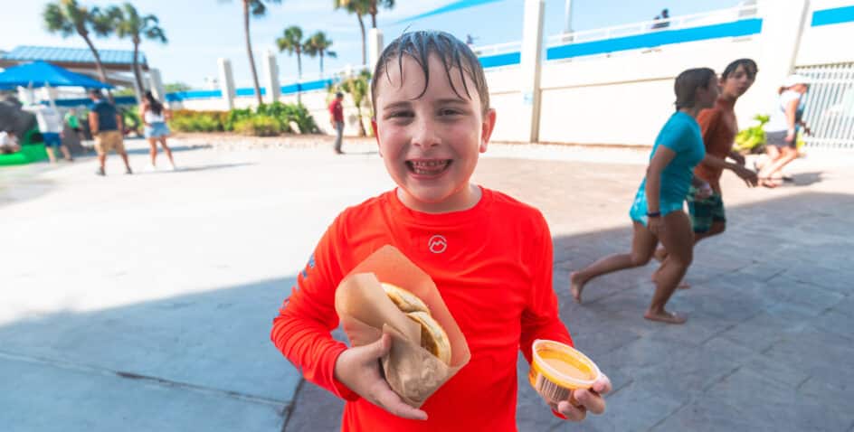 A smiling child in a red swim shirt, wet from playing, holds a sandwich and a cup of cheese sauce at an outdoor waterpark with people and palm trees in the background.