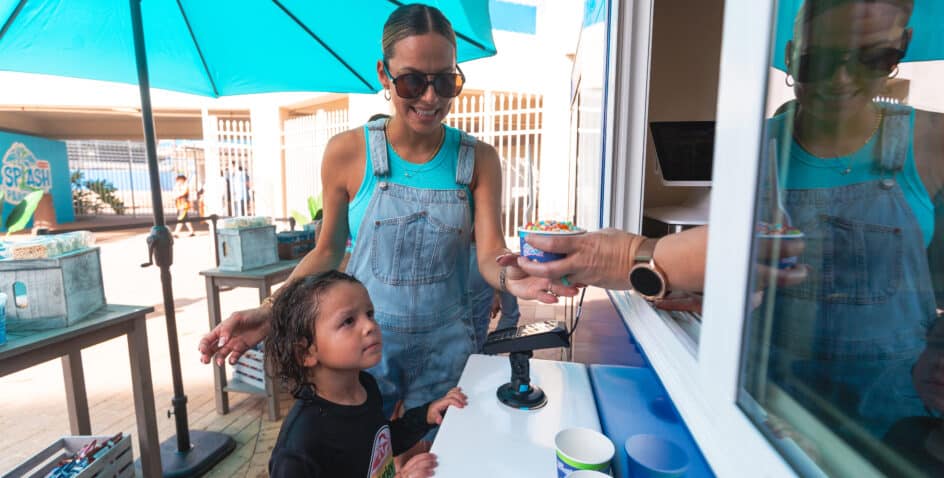 A woman in sunglasses and overalls receives a bowl of ice cream with sprinkles from a server at a window, while a young child stands beside her, looking up at the bowl.