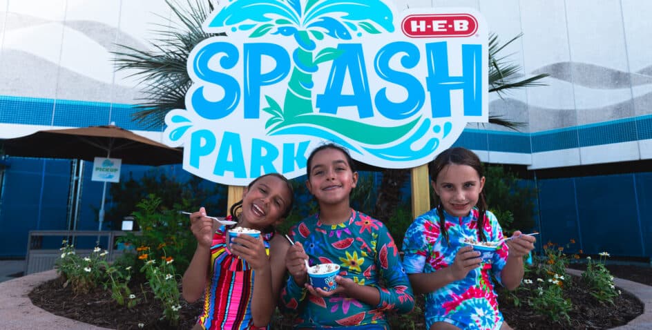 Three smiling children in colorful swimsuits eat ice cream in front of a sign that reads “HEB Splash Park,” with green plants and a building in the background.
