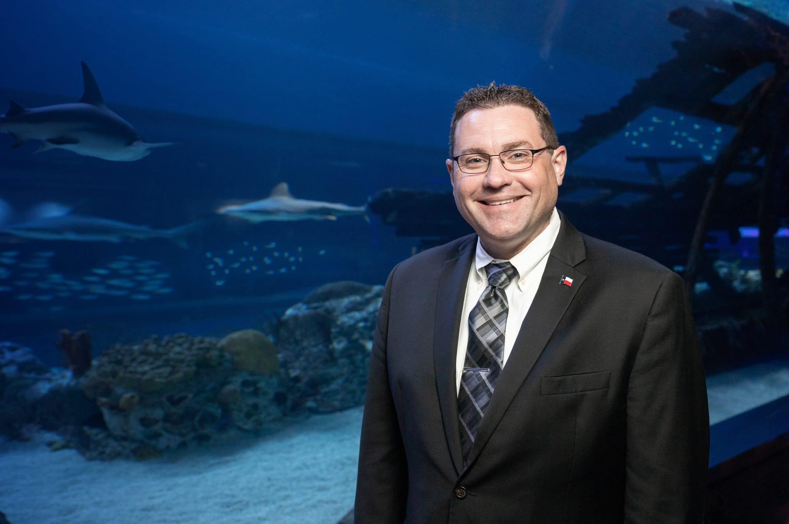 A man in a suit and tie smiles while standing inside an aquarium exhibit, with sharks swimming in a large tank and coral visible in the background.