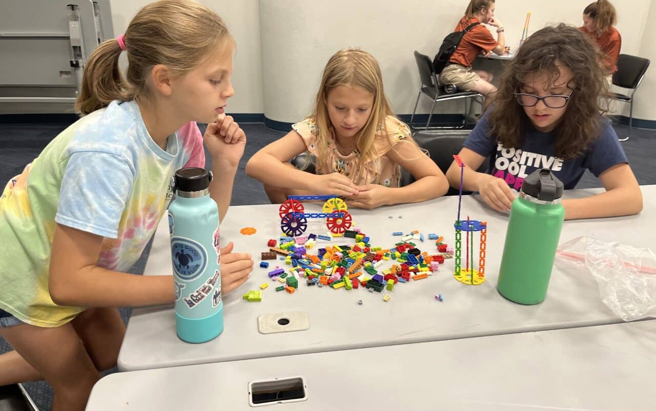 Three girls sit at a table building with colorful plastic blocks and gears. Two water bottles are on the table. More children are working in the background. The setting appears to be a classroom or activity room.