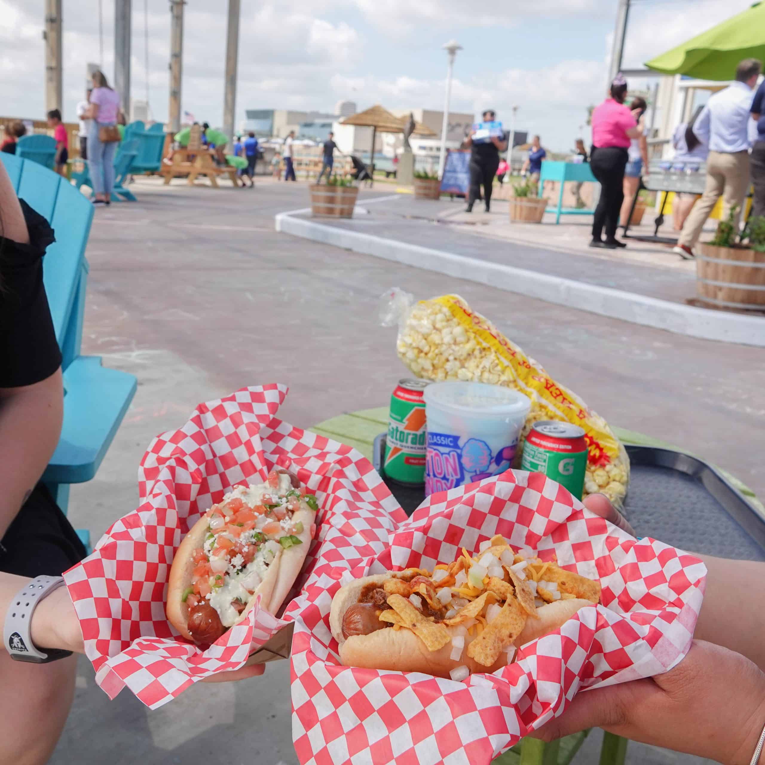 Two people hold hot dogs topped with various ingredients in checkered paper baskets at an outdoor seating area. A table in the background has canned drinks and a bag of popcorn, with people walking nearby.