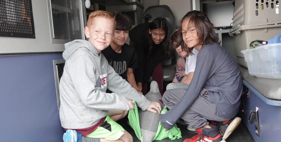 Five children are sitting inside a vehicle, smiling and gathered around a gray dummy or model. They appear to be participating in a hands-on activity or demonstration, with some kids touching or holding the model.