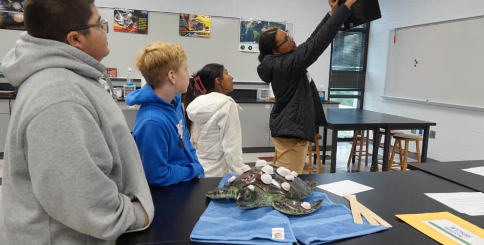 Four students in a science classroom observe as one student holds up a black object. On the table are two model turtles and papers. The classroom has lab tables, stools, and educational posters on the walls.