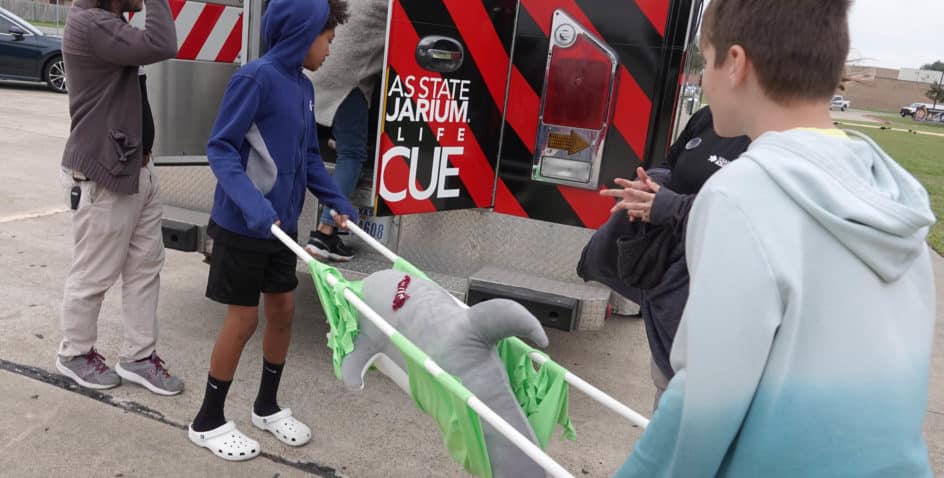 Three kids stand near an emergency vehicle, holding a stretcher with a large stuffed shark toy strapped in. One adult is partially visible, giving instructions. The scene appears to be an outdoor educational event.
