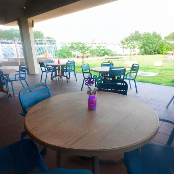 Outdoor seating area with round wooden tables and blue chairs on a tiled patio, overlooking a grassy lawn with trees. A jar holding pens and a decorative butterfly sits on the nearest table.