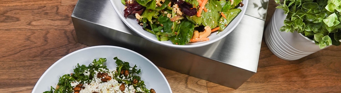Two bowls of fresh salad on a wooden table, one with leafy greens, carrots, and seeds, the other with greens, beans, and crumbled cheese. Two potted green plants are also visible in the background.