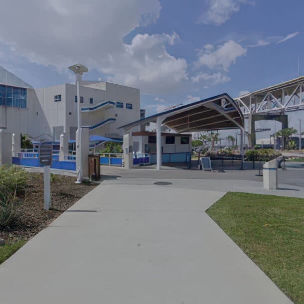 A modern outdoor walkway leads to a pavilion surrounded by landscaped greenery, palm trees, and large white buildings under a partly cloudy sky.