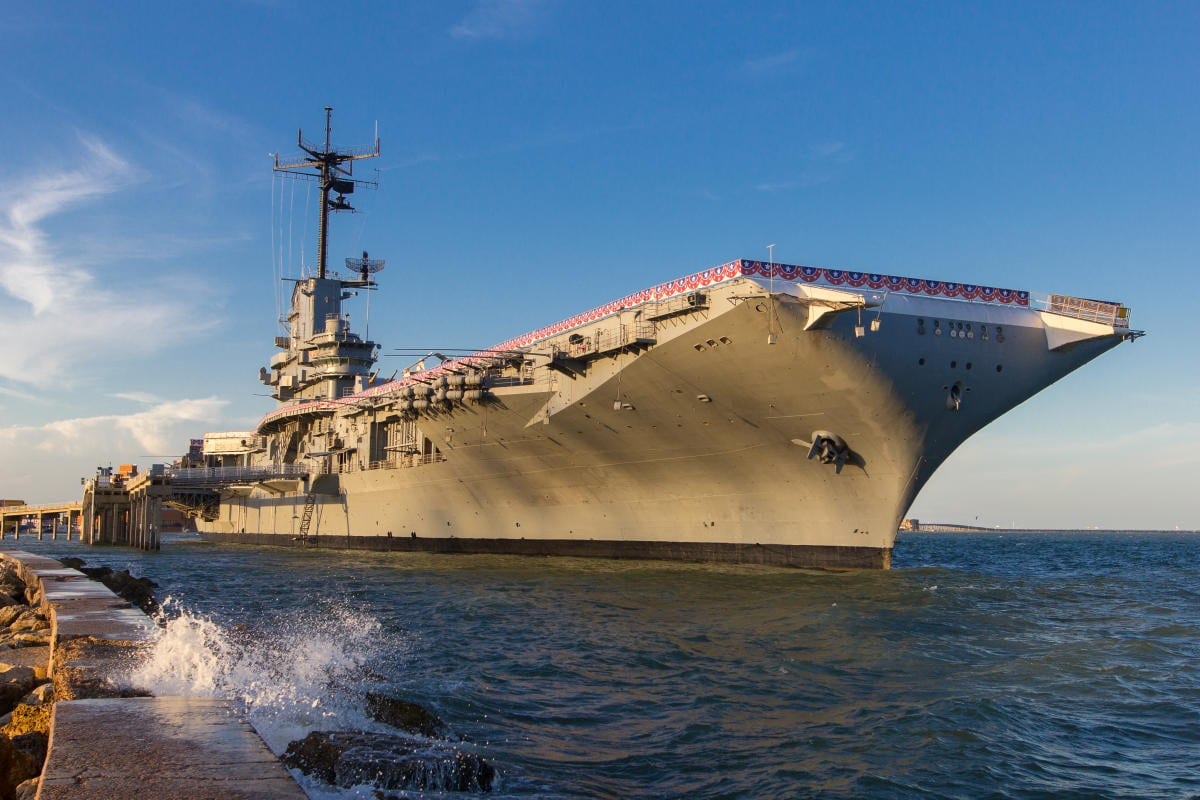 A large gray aircraft carrier decorated with red, white, and blue bunting is docked beside a pier, with gentle waves splashing against rocks in the foreground under a clear blue sky.