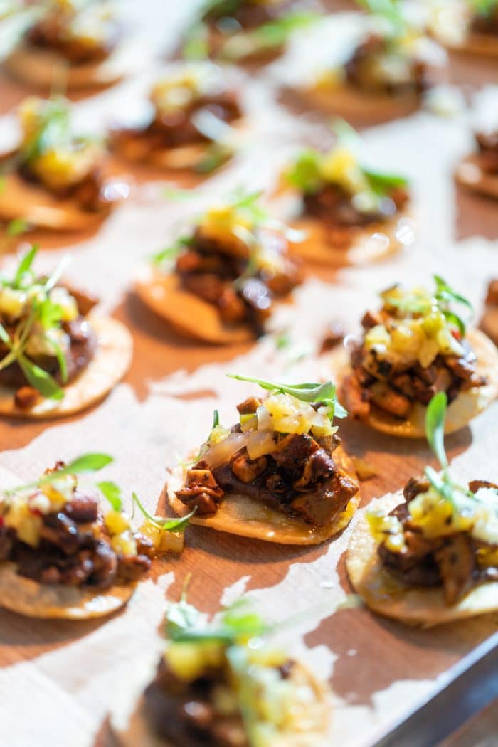 A close-up of small appetizer bites on round crackers, topped with cooked meat, diced vegetables, and garnished with fresh greens, arranged on a parchment-lined surface.