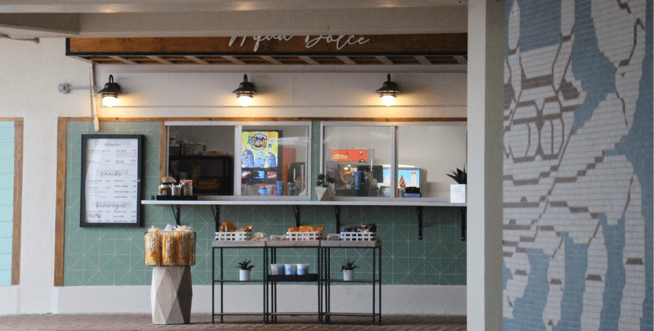 A small snack stand with a menu board, shelves of chips, and a counter with drinks, under three black lights. A large pixel-style mural is visible on the wall to the right.
