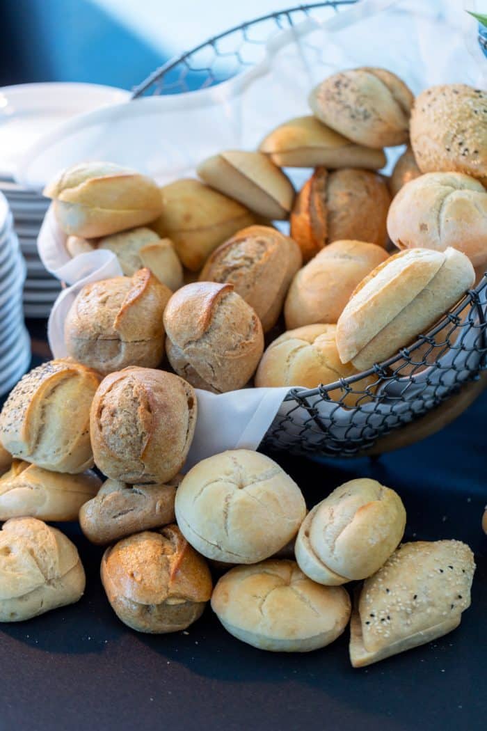 A wire basket lined with a white cloth is filled with assorted bread rolls, including plain, seeded, and poppy-seed varieties, with some rolls spilling onto a dark tabletop.