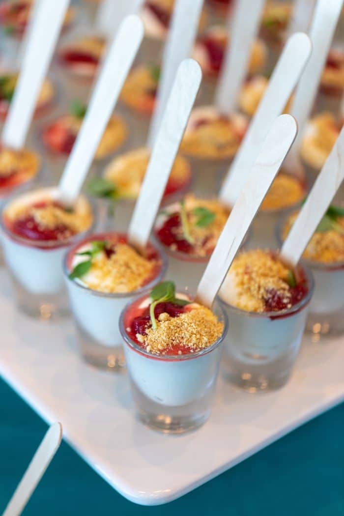 Mini dessert cups with layers of white cream, red fruit sauce, and crumbled topping, each served with a small wooden spoon and garnished with a mint leaf on a white tray.