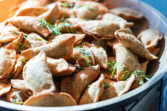 A close-up of a bowl filled with golden-brown, crispy fried dumplings garnished with fresh green herbs. The dumplings appear freshly made and are piled on top of each other.