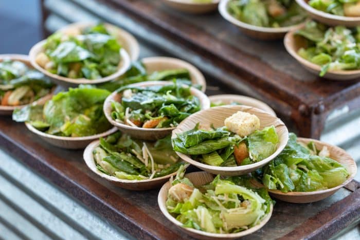 Small wooden bowls filled with fresh green salad, croutons, and grated cheese are arranged on rustic metal shelves, ready to be served.