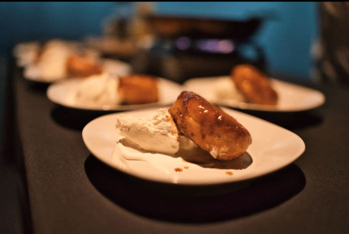 A close-up of a dessert plate featuring a golden-brown pastry with caramel sauce next to a generous scoop of vanilla ice cream, with similar plates blurred in the background.