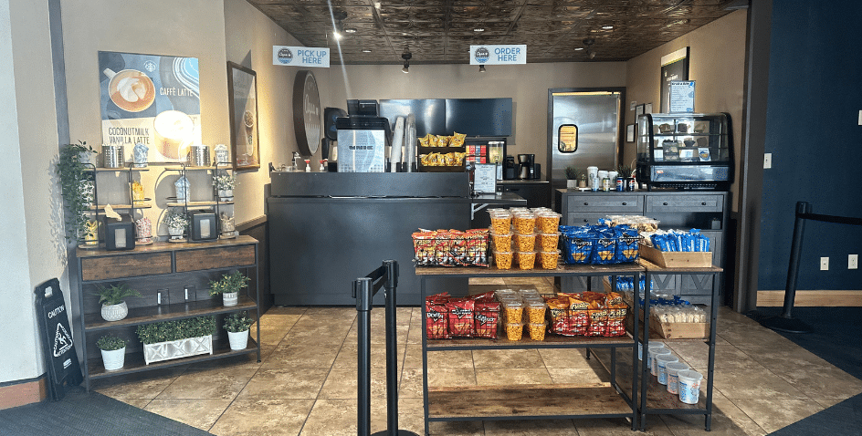 A small café counter with a snack display in front, offering chips, popcorn, and snacks. Behind the counter are coffee machines, pastries in a case, and signs for order and pickup. The area has a cozy, inviting setup.