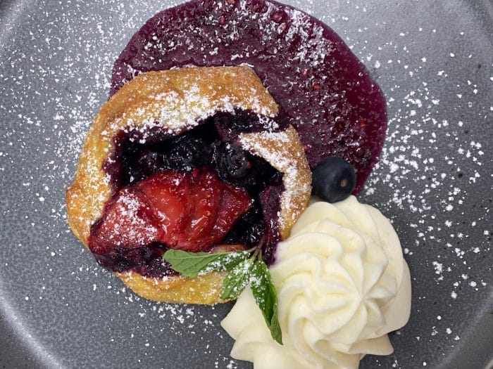 A small pastry filled with mixed berries sits on a gray plate, topped with powdered sugar, next to a swirl of whipped cream and garnished with a mint leaf. Berry sauce is spread underneath the pastry.