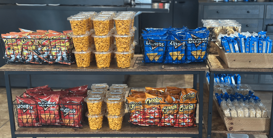 A display of assorted snacks on wooden shelves, including bags of Doritos, Lays, Cheetos, cookies, granola bars, and clear containers filled with goldfish crackers.