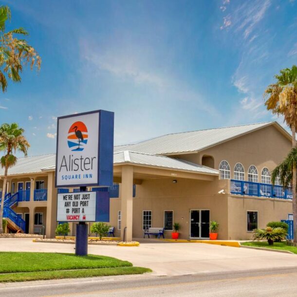 A two-story hotel named Alister Square Inn with palm trees out front and a large sign by the road. The building is tan with blue accents, under a sunny sky with scattered clouds.