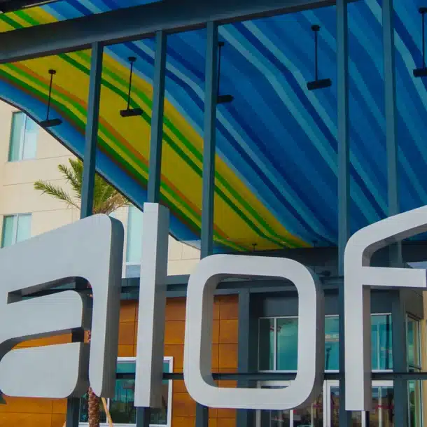 Modern hotel entrance with large silver aloft sign, colorful striped awning overhead, glass windows, palm trees, and brown paneling on the building façade.