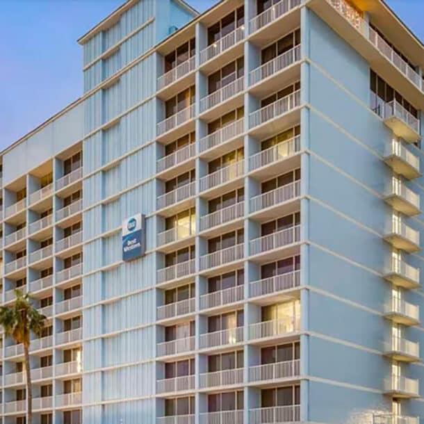 A multi-story light blue hotel building with balconies on each floor, illuminated by evening light. A single palm tree stands in front, and a hotel sign is visible near the middle of the building.