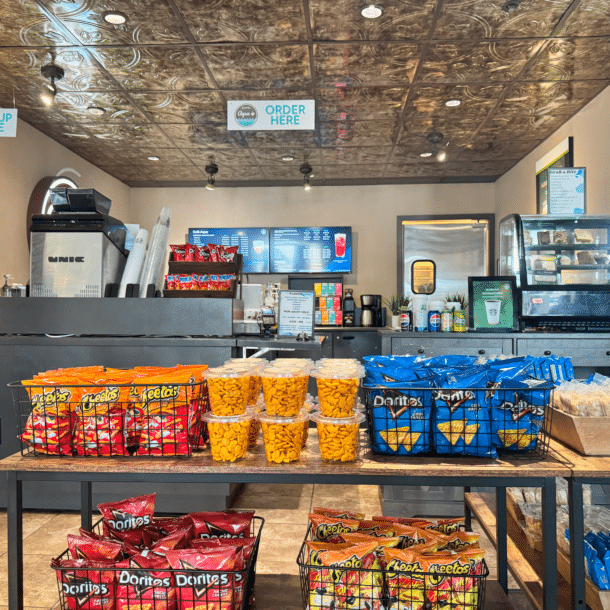 A snack counter with shelves displaying bags of Cheetos, Doritos, and other snacks. Behind the counter are drink machines, a glass food display, and signs reading “Pick Up Here” and “Order Here” hanging from the ceiling.
