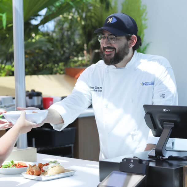 A chef in a white uniform and navy cap hands a bowl of food to a customer across a counter. The scene is bright and outdoors, with greenery in the background and plates of food on the counter.