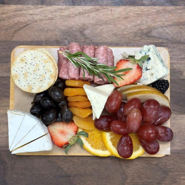 A charcuterie board with crackers, salami, rosemary, blue cheese, brie, black olives, dried apricots, grapes, orange slices, strawberries, and blackberries on a wooden surface.