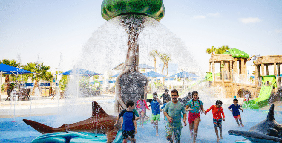 Children in swimsuits play under a large water fountain shaped like a tree in an outdoor water park, with slides, animal sculptures, and blue umbrellas in the background on a sunny day.