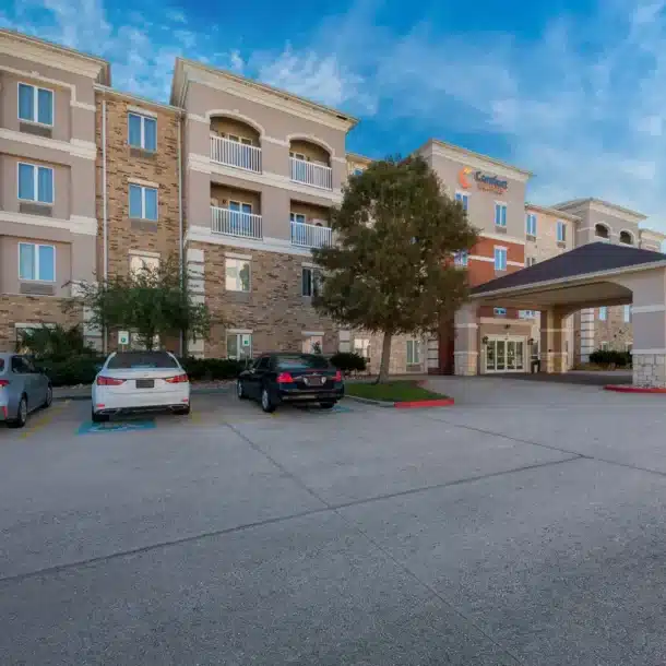 A modern, multi-story hotel with stone and stucco exterior, a covered entrance, three parked cars, and two flagpoles at the front, set against a bright blue sky with light clouds.
