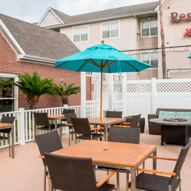 Outdoor patio area at a Residence Inn by Marriott with wooden tables, dark chairs, turquoise umbrellas, potted plants, and a nearby brick building, surrounded by a white fence.