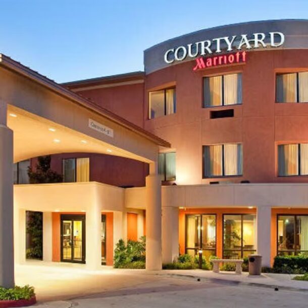 Exterior view of a Courtyard Marriott hotel with three stories, large entrance canopy, well-lit windows, landscaped greenery, and an American flag on the right. The address 5163 is displayed on the entrance canopy.