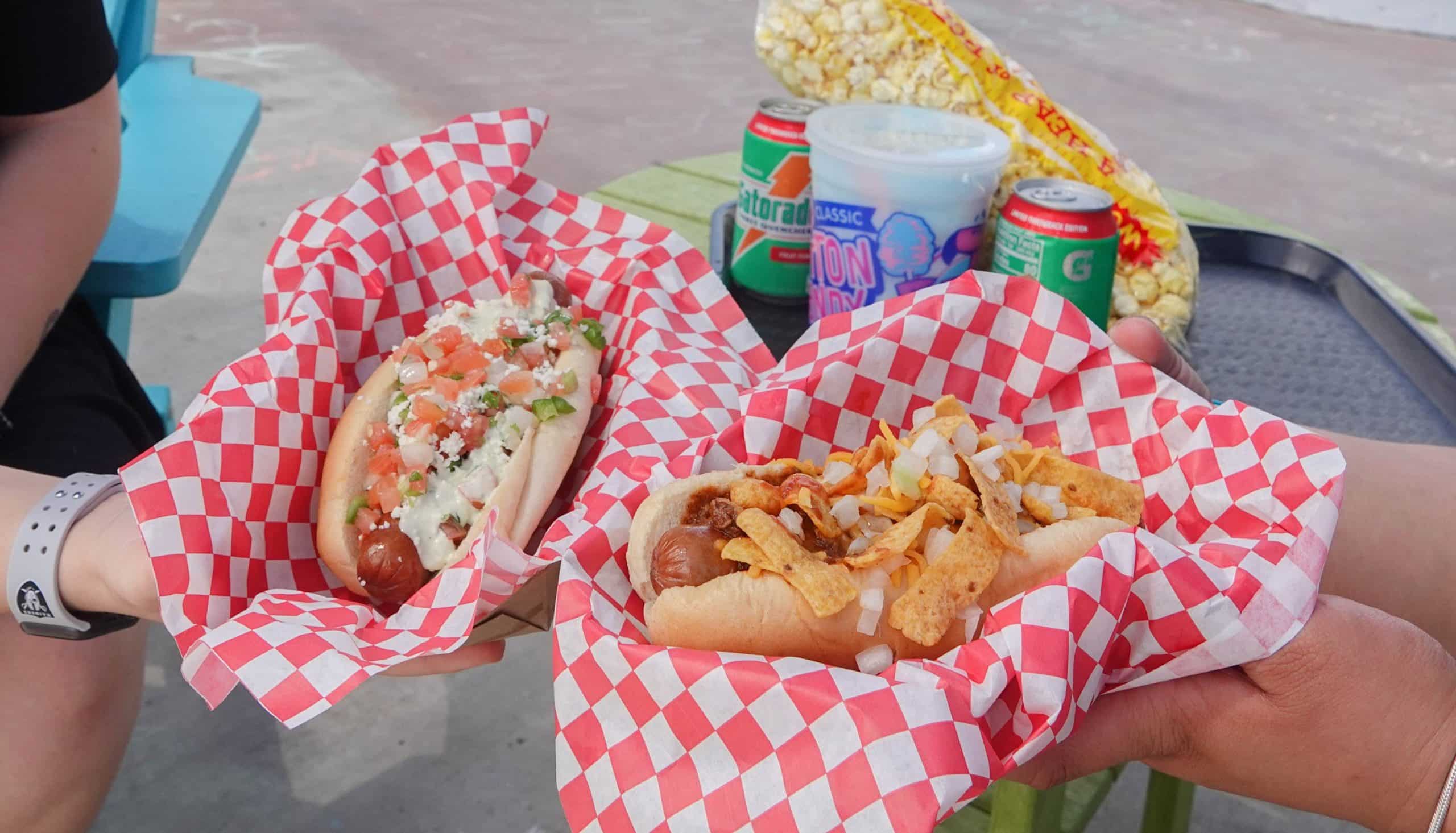 Two people hold hot dogs in red-and-white checkered baskets—one topped with pico de gallo, the other with corn chips and onions. On the table are popcorn, canned drinks, and a cup of blue slush.