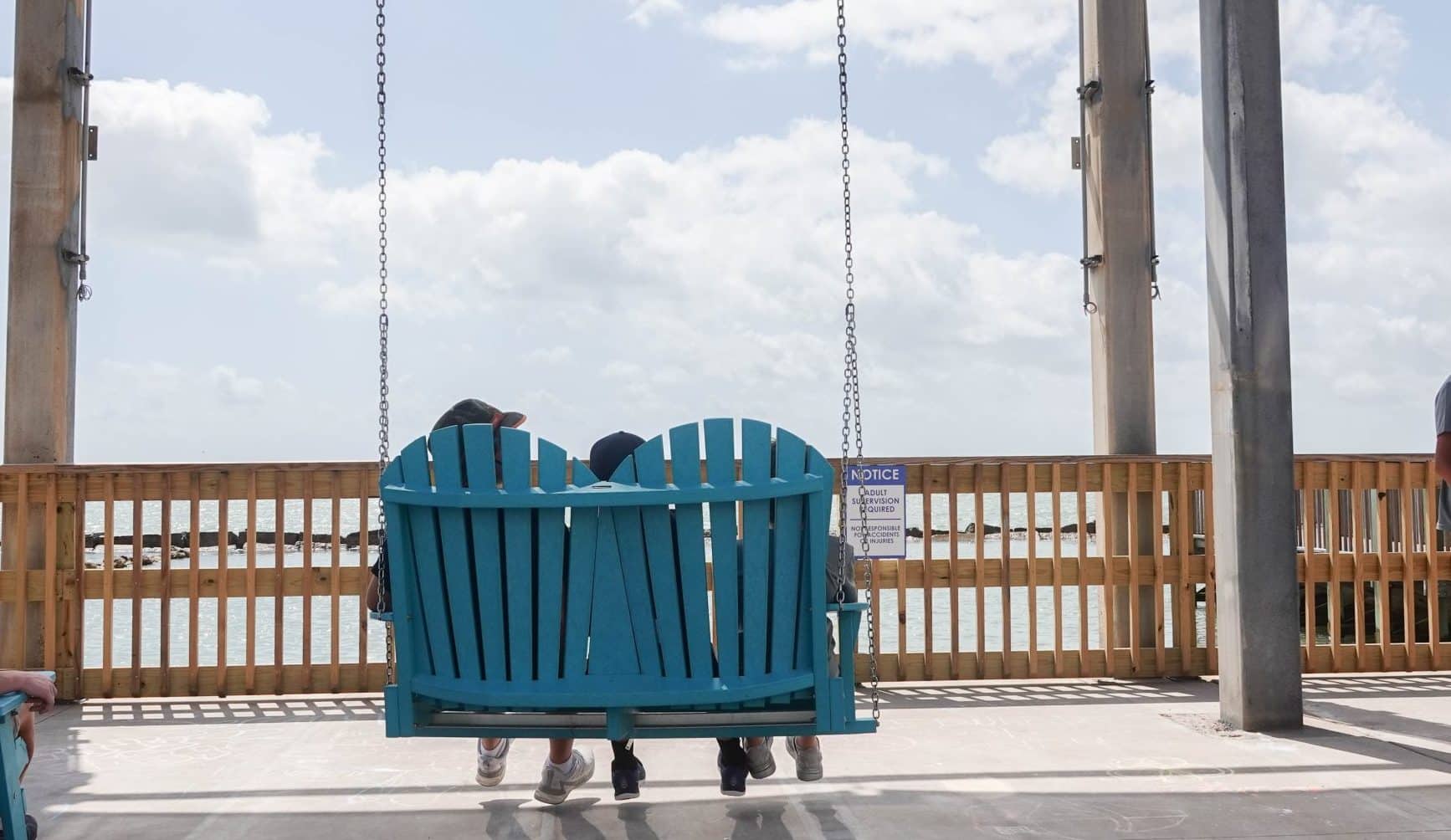Two people sit close together on a large blue swing facing the ocean, under a covered pier with wooden railings. The scene appears calm and sunny with a cloudy sky in the background.