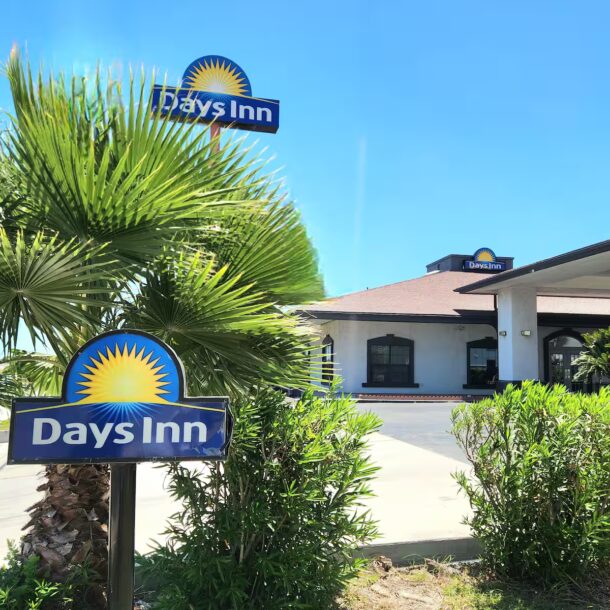 A Days Inn hotel exterior on a sunny day, with palm trees and bushes in front, and several Days Inn signs displaying the sunburst logo near the entrance.