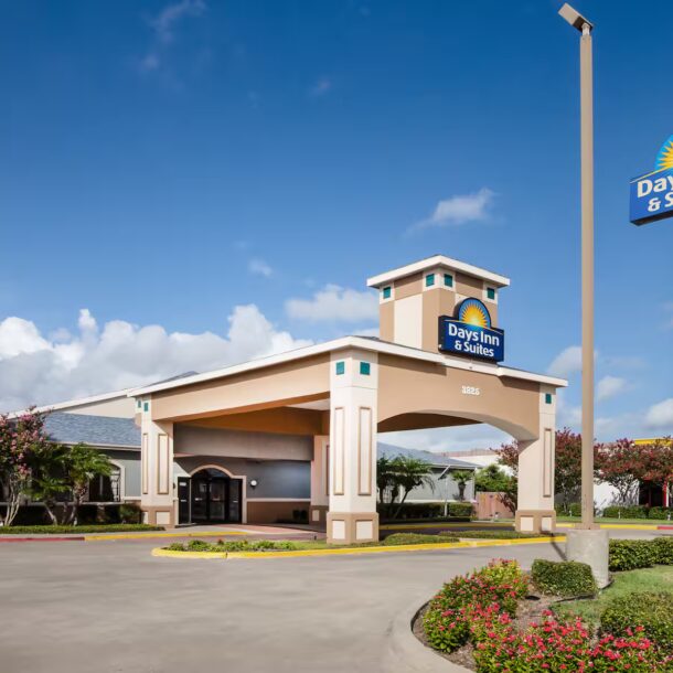 A Days Inn & Suites hotel exterior with a large entrance canopy, landscaped greenery, and visible signage, including a tall Days Inn sign and a Texas flag, under a partly cloudy sky.