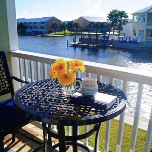 A small balcony overlooking a canal with waterfront houses. On the table are yellow flowers in a vase, an open book, and a mug. Two chairs sit beside the table in the sunlight.